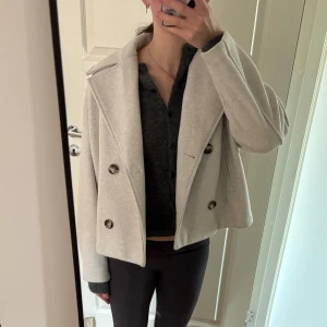 Beige kappa med knappar - Säljer en stilren beige kappa med stora knappar framtill. Jackan har en klassisk krage och är långärmad, perfekt för höst och vår. Den har en normal passform och ger en elegant look.