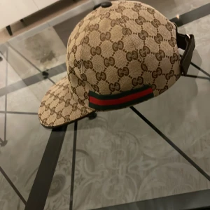 Beige keps med monogrammönster - Snygg beige keps från Gucci med det ikoniska monogrammönstret. Kepsen har en justerbar rem baktill och en klassisk skärm. Den har också de typiska gröna och röda ränderna på sidan.