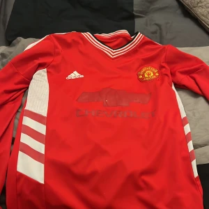 Röd fotbollströja från Adidas - Säljer en röd långärmad fotbollströja från Adidas med Manchester Uniteds emblem och Chevrolet-logga på framsidan. Tröjan har vita detaljer och Premier League-märke på ärmen. Perfekt för fotbollsfans! ⚽