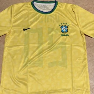 Gul Brasilien fotbollströja - Säljer en gul Brasilien fotbollströja från Nike med gröna detaljer runt halsen och det ikoniska brasilianska emblemet på bröstet. Perfekt för fotbollsfans som vill visa sitt stöd för landslaget. Tröjan har korta ärmar och en klassisk passform.