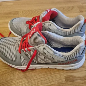 Grå och röda Reebok träningsskor - Reebok träningsskor i grått och rött med MemoryTech-sula för extra komfort. Skorna har ett ventilerande meshmaterial och röda snöre. Perfekta för träning eller en aktiv vardag.