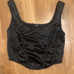 Svart korsettopp från bikbok - Snygg svart korsettopp från Reebok i glansigt material. Toppen har breda axelband och en stilren design som passar perfekt för en trendig look. Perfekt för att styla med både jeans och kjol.