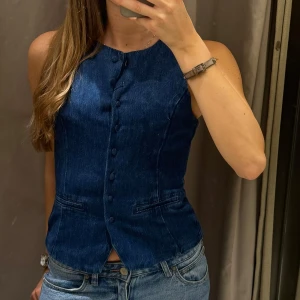 Mörkblå jeansväst - Snygg mörkblå jeansväst med knappar framtill. Västen har en figurnära passform och är perfekt för att ge en cool touch till din outfit. Den är ärmlös och har en klassisk krage.