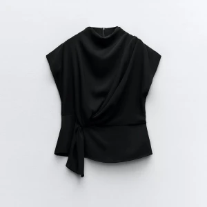 Svart draperad topp - Elegant svart topp med draperad design och knytning i sidan. Perfekt för en stilren look. Toppen har en unik form som ger en modern touch.