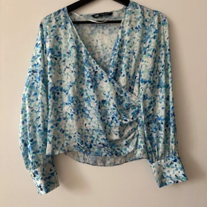 Mönstrad omlottblus från Zara - Säljer en snygg omlottblus från Zara med ett blått och vitt mönster. Blusen har långa ärmar med knappar vid ärmsluten och en v-ringad design. Perfekt för en stilren look!