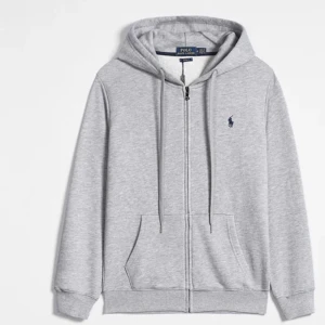 Grå hoodie från Polo Ralph Lauren - Säljer en stilren grå hoodie från Polo Ralph Lauren med dragkedja och det klassiska logotypbroderiet på bröstet. Tröjan har långa ärmar och en justerbar huva med snören. Perfekt för en avslappnad look.