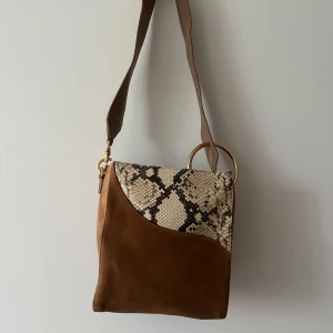 Brun axelväska med ormmönster från ATP Atelier - Snygg brun axelväska i mocka från ATP atelier med detaljer i ormmönster och guldfärgade ringar. Väskan har en bred axelrem och en unik design som kombinerar olika material och mönster. Axelremmen går att justera i längd. Väskan har även en dragkedja 