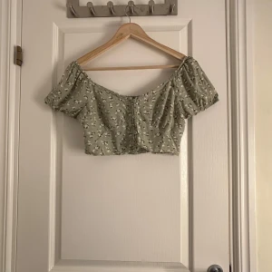 Grön blommig offshoulder topp från Hollister - Säljer en söt grön topp från Hollister med blommigt mönster. Toppen har korta puffärmar och en smockad baksida för en bekväm passform. Perfekt för varma dagar!