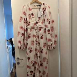 blommig långklänning från Gina Tricot - Elegant vit långklänning med blommigt mönster i rosa och vinröda toner från Gina Tricot. Klänningen har långa ärmar och en v-ringad design med knappar framtill. Perfekt för sommarens festligheter.                                                 Viral klänning från ginatricot och väldigt efterfrågad. Storlek 34💗