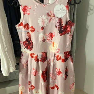 Blommig klänning från Happy Girls - Söt blommig klänning från Happy Girls i ljusrosa med röda och vita blommor. Klänningen är ärmlös och har knappar på baksidan. Perfekt för somriga dagar! Är i storlek 10/11 år för flickor☺️