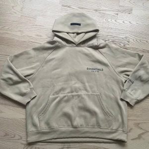 Beige hoodie från Essentials Fear of God - Säljer en stilren beige hoodie från Essentials Fear of God. Den har en klassisk känguruficka och ribbade muddar. Perfekt för en avslappnad stil med tryck på bröstet och ärmen.