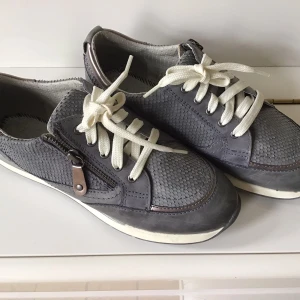 Grå sneakers med dragkedja - Snygga grå sneakers från Jana med vit sula och snörning. Skorna har en unik textur och praktisk dragkedja på sidan för enkel på- och avtagning. Perfekta för en stilren vardagslook.