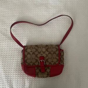Äkta coach väska - Snygg axelväska från Coach i beige med röda detaljer och ett klassiskt mönster. Väskan har en justerbar axelrem och ett spänne framtill. Perfekt för att ge din outfit en stilfull touch. Det är en äkta coach väska! Man kan multistyla den genom att göra handtaget mindre eller längre genom att vika den inuti väskan. Om du behöver fler bilder hör gärna av dig 🩷