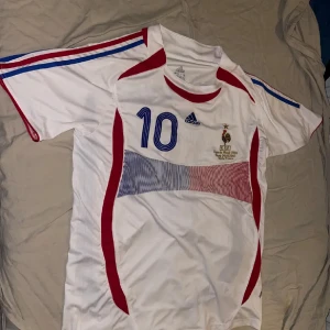Vit fotbollströja från Adidas - Säljer en vit fotbollströja från Adidas med blå och röda detaljer. Tröjan har nummer 10 och text på ryggen. Perfekt för fotbollsfans som vill ha en klassisk look.