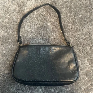 Svart axelväska med krokodilmönster - Snygg svart axelväska med krokodilmönster och dragkedja. Perfekt för att ge en stilren touch till din outfit. Väskan har en justerbar axelrem och är tillverkad i syntetmaterial.