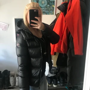 Svart dunjacka från Colmar - Säljer en snygg svart dunjacka från Colmar. Jackan har en pufferdesign med dragkedja och en praktisk huva. Perfekt för kyliga dagar!