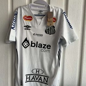 Vit fotbollströja från Umbro - Säljer en vit fotbollströja från Umbro med flera sponsorer tryckta på framsidan och baksidan. Tröjan har korta ärmar och ett tryck med nummer 10 och texten 'Neymar Jr' på baksidan. Perfekt för fotbollsfans!