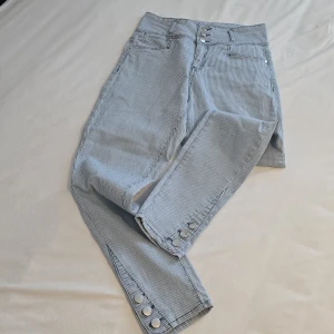 Randiga jeansbyxor med knappar - Snygga randiga jeansbyxor i ljusblått och vitt med hög midja. Byxorna har en rak passform och är dekorerade med knappar vid bensluten. Perfekta för en stilren look!