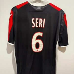 Seri - Snygg svart fotbollströja från OGC Nice med röd och vit detaljering. Tröjan har korta ärmar och spelarens namn 'Seri' samt nummer 6 på ryggen. Perfekt för fotbollsfans som vill visa sitt stöd för laget.   Skriv gärna för fler bilder