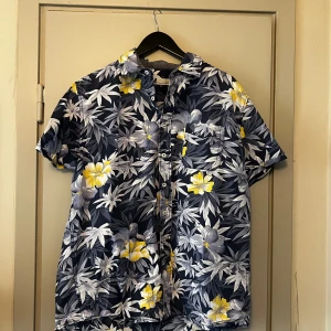 Blommig kortärmad skjorta - Säljer en snygg blommig skjorta från dressman i storlek XL. Skjortan har ett tropiskt mönster med gula och vita blommor på en mörkblå bakgrund. 