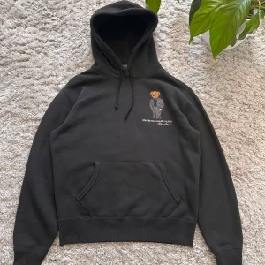 Svart hoodie från Ralph Lauren med nallebroderi - Säljer en svart hoodie från Ralph Lauren med ett coolt broderi av en nalle på framsidan. Hoodien har en klassisk känguruficka och justerbar huva med dragsko. Perfekt för en avslappnad stil! Detta var AI beskriving så fråga gärna mig om du är intresserad.