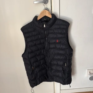 Svart dunväst från Polo Ralph Lauren - Snygg svart dunväst från Polo Ralph Lauren med dragkedja och två fickor framtill. Perfekt för lager-på-lager under kyliga dagar. Den har en diskret logga på bröstet och en stilren design.