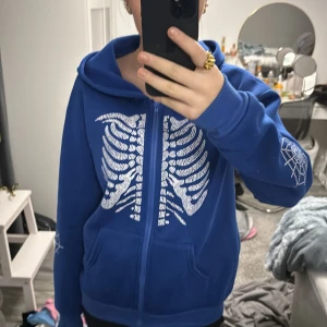 Blå hoodie med skelettmönster från Shein - Cool blå hoodie från Shein med dragkedja och ett unikt skelettmönster på framsidan. Ärmarna har ett spindelnätsmönster som ger en extra touch. Perfekt för dig som vill sticka ut!