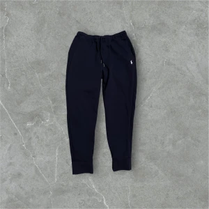 Mörkblåa mjukisbyxor från Polo Ralph Lauren - Säljer ett par svarta mjukisbyxor från Polo Ralph Lauren med en liten broderad logga på sidan. Byxorna har en bekväm passform med snörning i midjan och ribbade muddar vid bensluten.