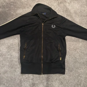 Vintage Fred Perry track jacket - Snygg svart jacka från Fred Perry med dragkedja och vita randdetaljer längs ärmarna. Jackan har två fickor med dragkedja och ett broderat märke på bröstet. Perfekt för en stilren look. Zippen e trasig men funkar ändå