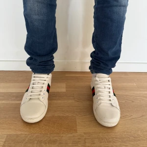 Gucci skor - Snygga vita sneakers från Gucci med klassiska röd-gröna ränder och broderad detalj på sidan. Skorna har snörning och en unik design med röd och grön häl. Perfekta för att ge din outfit en lyxig touch. Priset är väldigt diskuterbart och skicket är 10/10