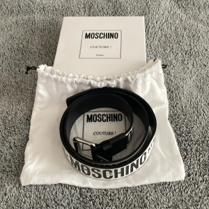 Svart och vit bälte från Moschino - Snyggt svart och vitt bälte från Moschino med tydlig logotyp. Bältet har en klassisk design med ett silverfärgat spänne. Perfekt för att ge en stilren touch till din outfit.