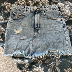 Blå jeanskjol från Pull&Bear - Snygg blå jeanskjol från Pull&Bear med slitna detaljer och fransig nederkant. Perfekt för en avslappnad stil. Kjolen har klassisk femficksdesign och knappgylf.