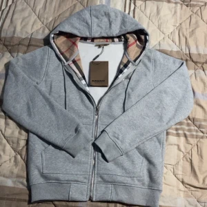 Grå hoodie från Burberry - Säljer en stilren grå hoodie från Burberry med dragkedja och klassiskt rutigt mönster i huvan. Tröjan har en broderad logga på bröstet och är tillverkad i mjuk bomull med lite elastan för extra komfort. Perfekt för en avslappnad look.