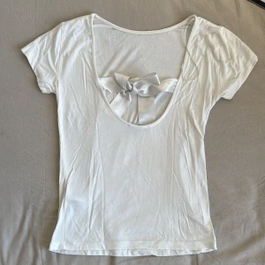 Vit kortärmad topp med rosett - Säljer en söt vit kortärmad topp med en stor rosett i ryggen. Toppen har en djup urringning bak som ger en fin detalj. Perfekt för en somrig look!