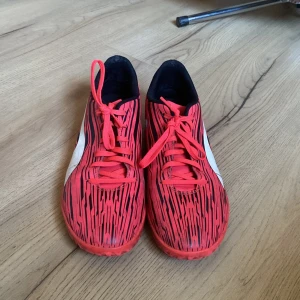 Röda fotbollsskor från Puma - Säljer ett par röda fotbollsskor från Puma med svart mönster och vita detaljer. Skorna har snörning och är perfekta för inomhusspel. De har en modern design med en bekväm passform.