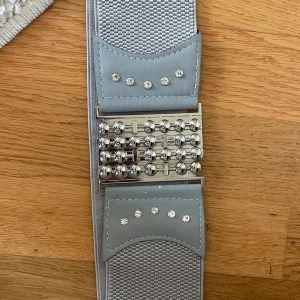 Grått elastiskt bälte med strassdetaljer - Snyggt grått bälte med elastiskt material och glittriga strassdetaljer. Bältet har en unik design med silverfärgat spänne och dekorativa nitar. Perfekt för att ge en extra touch till din outfit.
