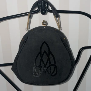 Svart handväska med kedjedetaljer - Elegant svart handväska med guldfärgade kedjedetaljer och en unik broderad design på framsidan. Väskan har en klassisk stängning upptill och en stilren form som passar perfekt för en chic look.