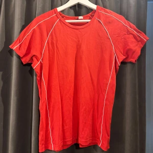 Röd t-shirt i träningsmaterial  - Säljer en röd t-shirt med vita sömmar längs ärmarna och sidorna, passar för träning men även för vardagen. Den har en rund halsringning och korta ärmar. Skriv vid frågor och innan köp! 