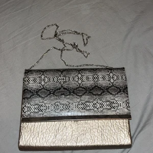 Elegant kuvertväska med ormmönster - Snygg kuvertväska med en mix av ormmönster och guldiga detaljer. Väskan har en kedjerem och stängs med en tryckknapp. Perfekt för att ge en touch av glamour till din outfit.