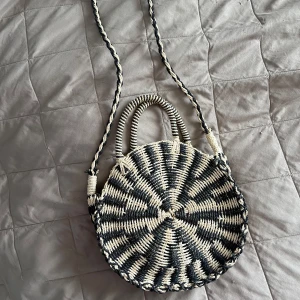 Rund axelväska i flätad stil - Säljer en rund axelväska i svart och beige med flätat mönster. Väskan har både axelrem och handtag, samt innerfack med dragkedja. Perfekt för dig som vill ha en unik accessoar till din outfit!