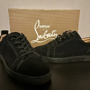 Svarta sneakers från Christian Louboutin - Säljer ett par svarta sneakers från Christian Louboutin med klassisk röd sula. Skorna har snörning och är tillverkade i mocka med svarta detaljer. Perfekta för dig som vill ha en stilren och lyxig look.