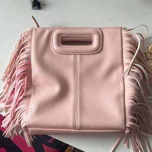 Ljusrosa axelväska med fransar - Säljer en maje liknande väska med fransar på sidorna och inbyggda handtag. Väskan har ett stilrent och modernt utseende, tillverkad i syntetmaterial med struktur. Perfekt för dig som vill ha en unik accessoar med lite extra detaljer. Den har några mörkare rosa fläckar på fransarna, jag vet inte hur den kom dit. De är från en tidigare köpare, men det är inget som märks när man bär den😊