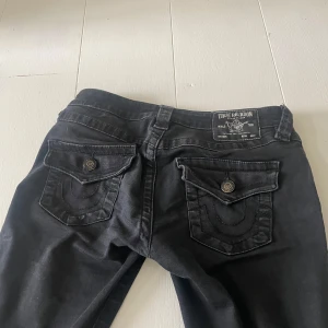 Svarta jeansbyxor från True Religion Joey - Säljer ett par svarta True Religion Joey jeans med låg midja och flare-ben. Dem passar mig som är 165. Inga mer defekter förutom att knapparna har tappat färg som man ser på bilderna.                Skriv för mer frågor och bilder💕