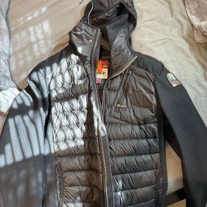 Svart hybridjacka från Parajumpers - Säljer en svart hybridjacka från Parajumpers med quiltade partier framtill och på ryggen, samt släta ärmar och sidor. Jackan har huva, dragkedja och en bröstficka med dragkedja. Perfekt för lager-på-lager.