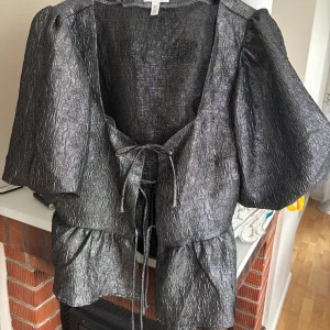 Glittrig silvrig blus med puffärm från H&M - Supersnygg silvrig blus från H&M med glittrig finish och puffiga korta ärmar. Blusen har knytning framtill och volang nedtill, perfekt för att sticka ut. Materialet är syntet och ger en skimrande look.