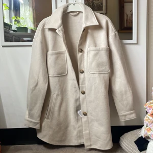 Beige kappa från Vila - Säljer en beige overshirt från Vila med stora bröstfickor och knappar framtill. Jackan har lång ärm och en rak, avslappnad passform. Perfekt att slänga över en hoodie eller t-shirt för en snygg lager-på-lager-look. Kappan är vad jag tror i storlek L, aldrig använd