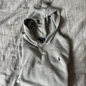 Grå hoodie från Polo Ralph Lauren - Säljer en klassisk grå hoodie från Polo Ralph Lauren med broderad logga på bröstet. Tröjan har huva, magficka och ribbade muddar vid ärmslut och nederkant. Perfekt för en avslappnad stil.