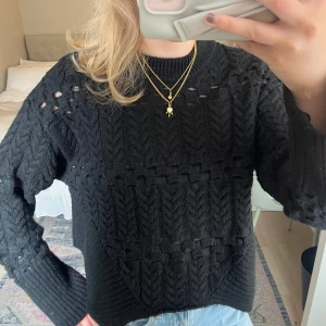 Svart stickad tröja från Zara - Säljer en svart stickad tröja från Zara med snyggt hålmönster och breda ribbade muddar. Tröjan har lång ärm och rund halsringning. Perfekt till jeans eller kjol för en trendig look.