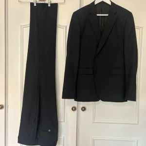 Svart kostym från Bläck - Klassisk svart kostym från Bläck. Kavaj i modell Neptune Sharkskin. Regular fit. Kavaj med två knappar och byxor med knappstängning. Perfekt för formella tillfällen. 