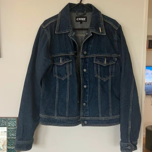 Mörkblå jeansjacka från CHNK - Säljer en klassisk mörkblå jeansjacka från CHNK med knappar framtill och två bröstfickor med lock. Jackan har en rak passform och tydliga sömdetaljer. Perfekt att slänga över en t-shirt eller hoodie.
